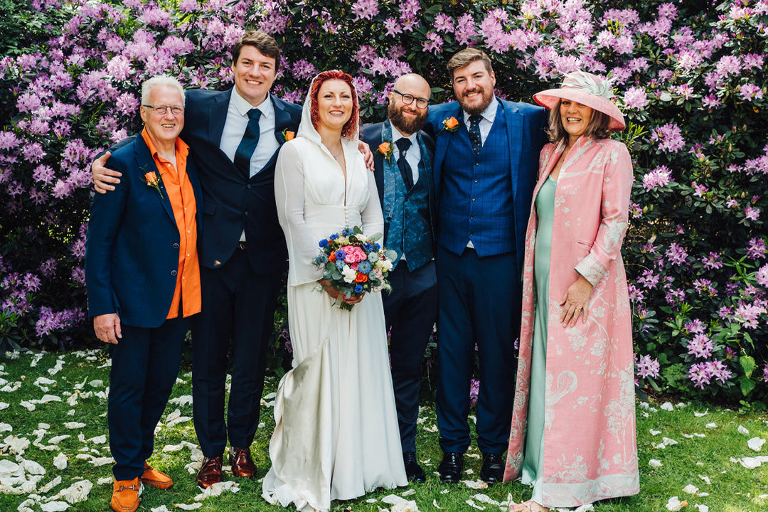 Mother of the bride wearing a stunning Shibumi outfit for her daughter wedding. Long cashmere pink coat for mother of the bride. The family is standing in front of purple blossoms. 