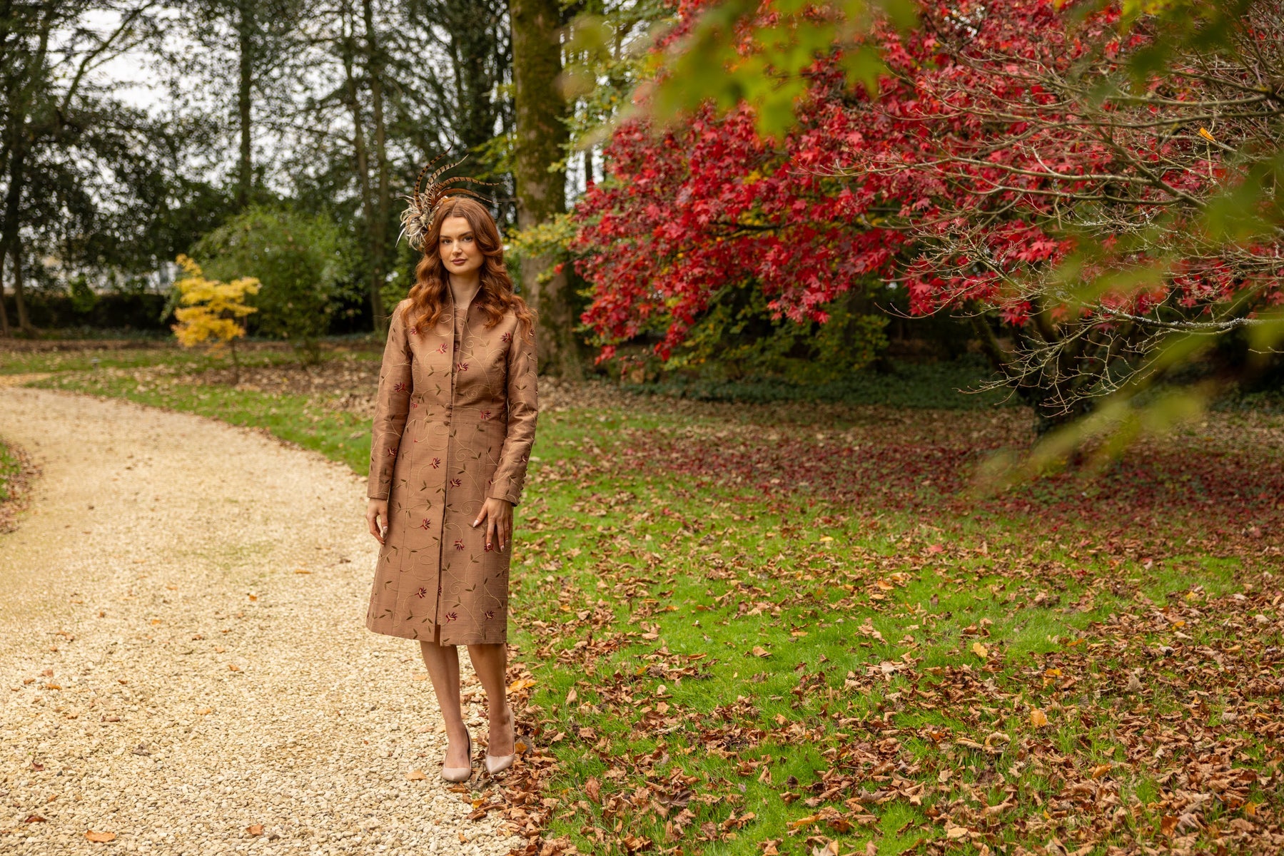 Dark haired woman stands in a garden in a red dress and green embroidered silk coat, the Avani coat by Shibumi.