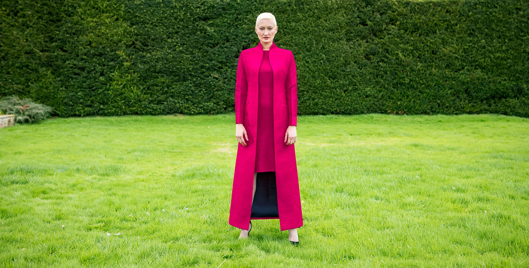 A woman with short blonde hair in a bright pink Hepburn silk dress and floor length Devi silk coat.