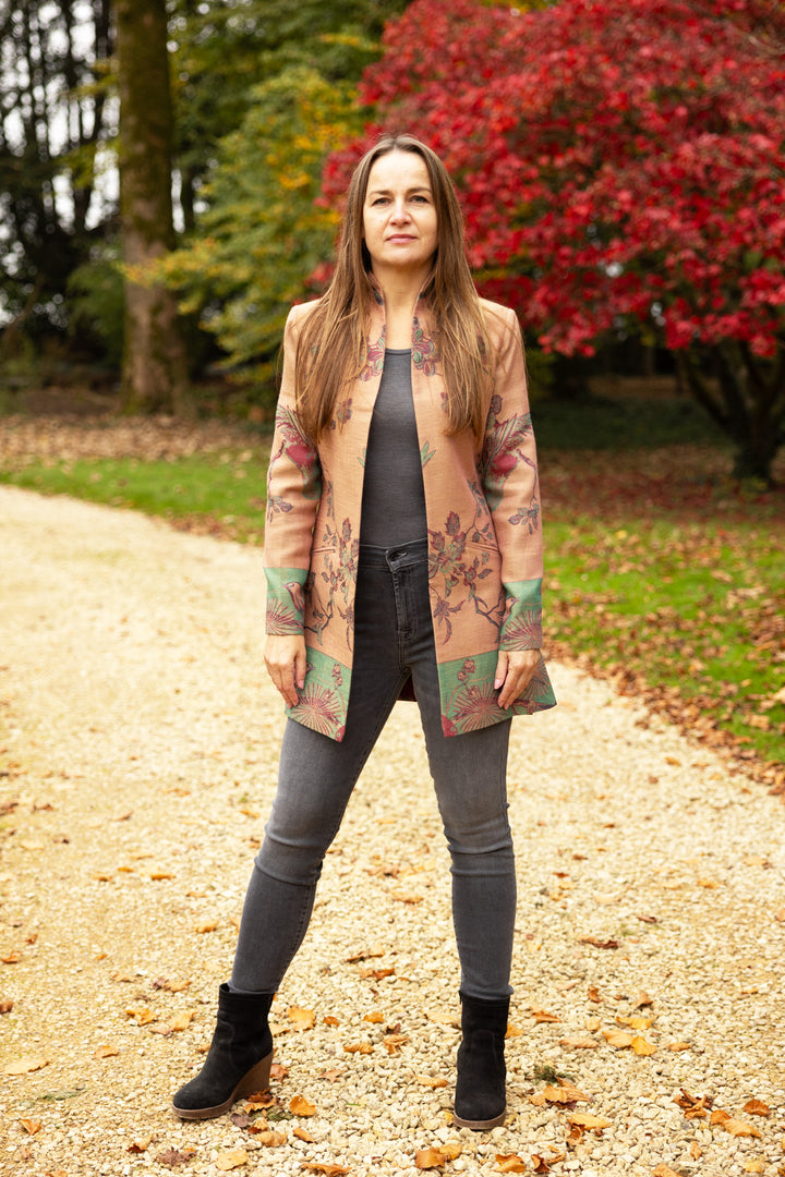 Lady standing in front of trees with autumn red leaves.  Wearing a very cool and unique jacket with jeans. Pink jacket with green, red and purple flowers. This is a jacket that can be worn smart or casual. 