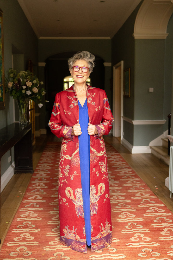 Mature lady wearing standing in a hallway wearing a striking maxi coat. Long red coat, with yellow and purple pattern, birds and flowers. The coat is worn open with blue silk kaftan underneath. 