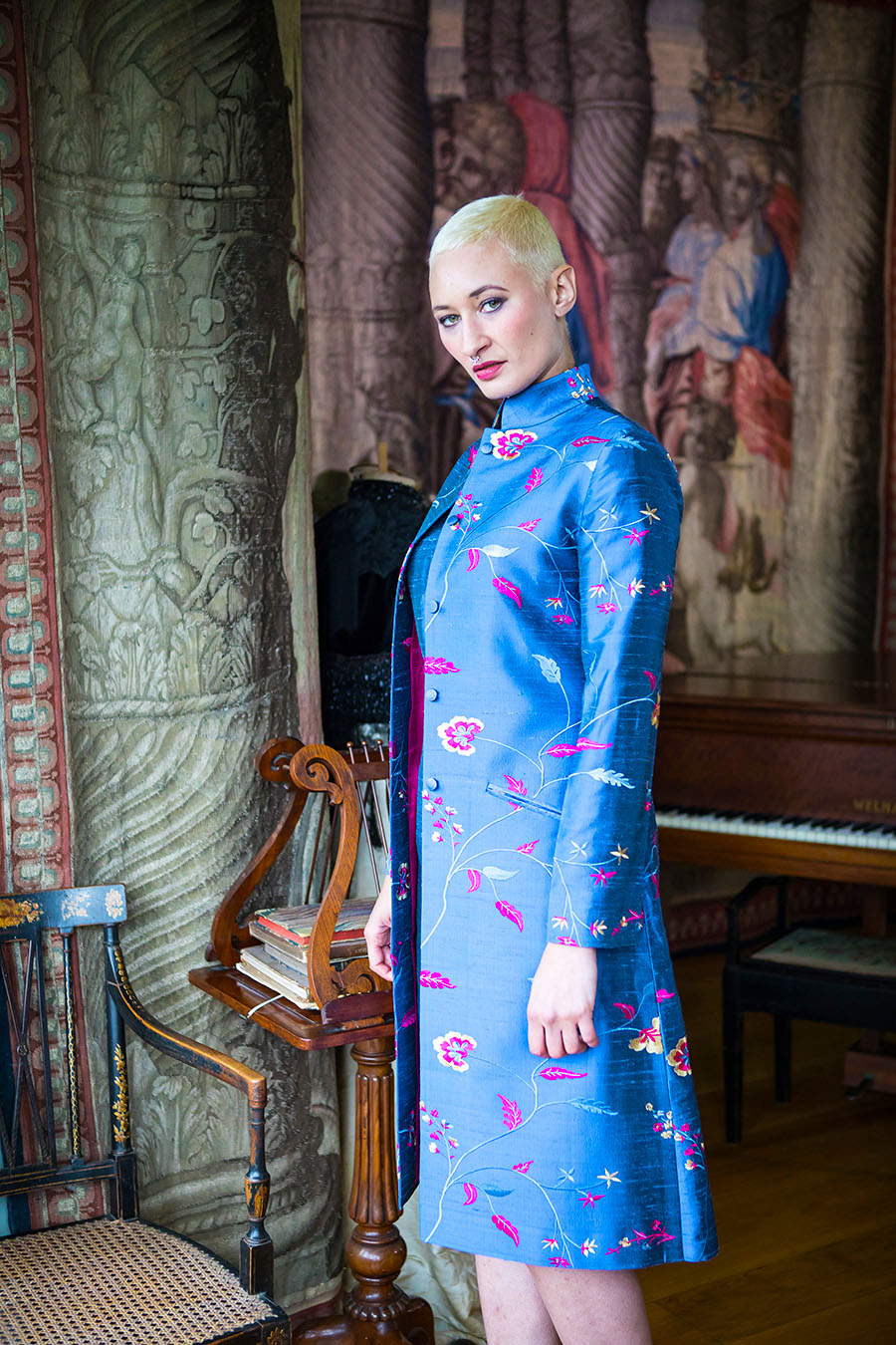 Woman standing to the side in a smart nehru frock coat with a mandarin collar and slit pocket, in a blue embroidered raw silk with a lighter blue, pink and yellow pattern